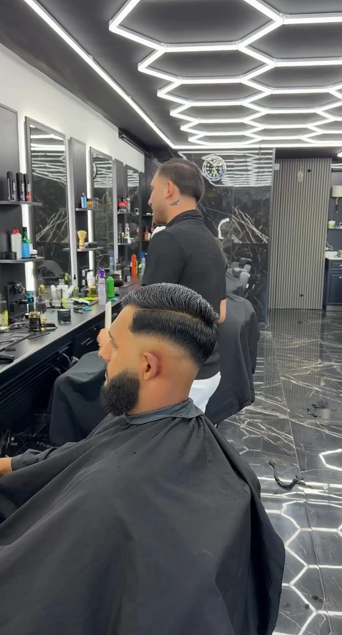 Barber aan het werk achter een klant met een verse fade en strakke baard, in de zaak met het lichtplafond